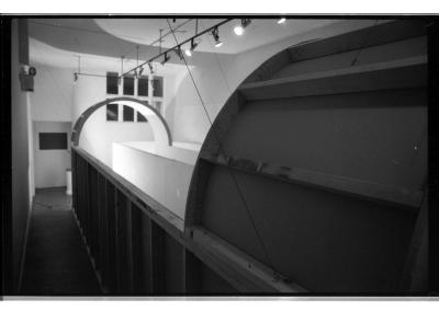 Seen in black and white, an architectural structure made of white wooden planks stands inside a gallery. Seen from the outside of the structure, two arched walkways cover a winding, narrow walkway.