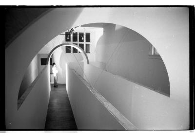 In a gallery, a person stands at the arched entrance of an architectural structure built from two arches and a winding, narrow walkway between. The gallery’s high-angle lighting illuminates the figure, with their shadow enlarged on the back wall. The image is in black and white.
