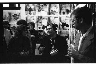 A group of people including Cornelia Wyngaarden, Herman Nitsch and Vincent Trasov stand in Western Front’s gallery, where a number of large-scale photos documenting the ritual mutilation of a nude body are tiled on the walls.