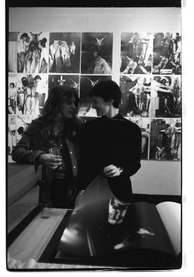 Cornelia Wyngaarden and Jane Ellison stand close to each other while in conversation. Ellison flips a page of a large-scale photo book, and Wyngaarden holds a glass in her right hand. Behind them is a photo installation showing documentation of the ritual mutilation of a nude body. 