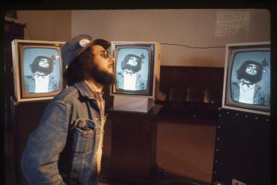 Michael Goldberg stands in front of three monitors displaying his image in the Grand Luxe Hall. He wears a denim jacket and hat and faces the camera in profile. 