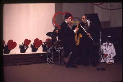 Wearing a tuxedo, Eric Metcalfe plays a handmade saxophone painted with leopard spots. He is joined on the stage of the Grand Luxe Hall by Taki Bluesinger on drums and Don Druick on flute. The back of the room is decorated with paper cutouts of black hand shapes holding red hearts.