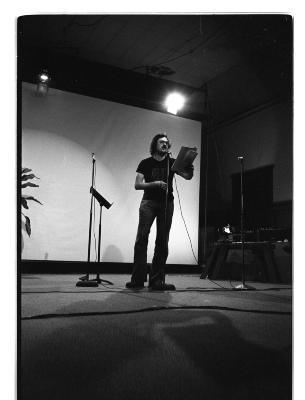 One person stands alone at a microphone. There is one microphone on a stand to either side of them. They stand in front of a blank projector screen. The person is back lit by a spotlight, their shadow lays in front of them. 