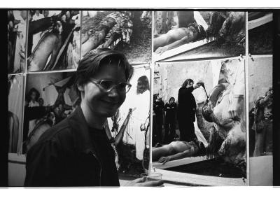 A male presenting person with short hair and glasses stands in front of a photographic installation by Hermann Nitsch that shows the ritual mutilation of a nude body.