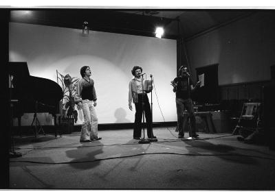 Three people stand singing in front of two microphones, using hand and gestures and wearing excited expressions. Two people are holding notebooks. Behind the trio there is a grand piano and a plant. The three are illuminated by a spotlight. 

