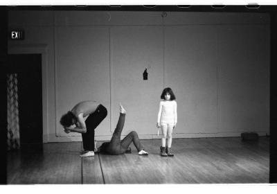 Lola Ryan stands in a forward fold with bent knees, cradling her head in her hands while Jane Ellison lies on the floor with one leg extended towards the ceiling. A child dressed in a long sleeve shirt, shorts, and combat boots stands next to them. The image is a black-and-white photograph.