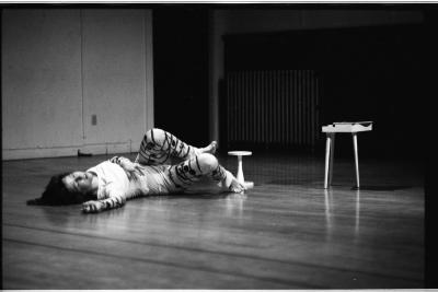 A dancer lies on the floor with one of their legs bent in the air and their right arm sprawled out. The dance studio around them has wooden floors, walls with wainscoting, and a radiator in the back. 