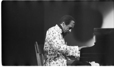 A medium-long shot of a man performing as he plays the grand piano. There is a spotlight on him and he is wearing a light-coloured patterned blazer. 
