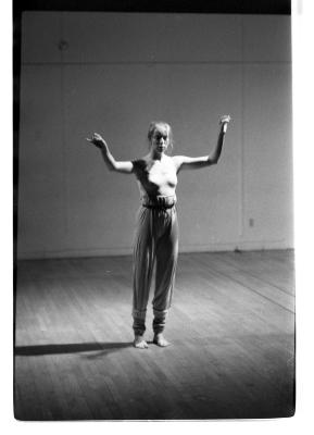 A woman stands in a dance studio, her arms loosely raised up in the air. With her right hand, she points her index and pinky finger extended. Her left hand is open in the air. She is wearing a tank-top and long pants. The image is black and white.