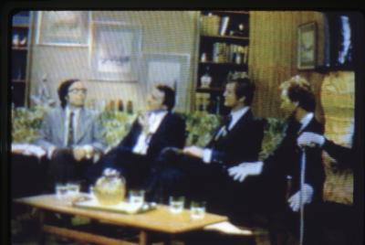 A television broadcast of four men in suits, including Art Phillips and John Mitchell, and Mr. Peanut sitting on a couch in a living-room setting. 