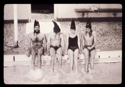 Jorge Zontal, Kate Craig, an unnamed subject, and Glenn Lewis sit on the edge of the pool deck with their feet splashing in the water at Crystal Pool. They are all wearing bathing suits and bathing caps that resemble shark fins. 
