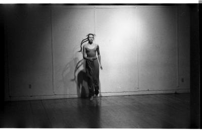 A woman jumps in a dance studio. She is wearing a tank-top, long pants, and her hair is in two long braids. The image is black and white.
