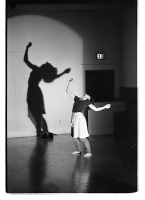 A black-and-white photo of Jane Ellison bending backwards with her arms outstretched. She is illuminated by a spotlight that projects her shadow on the back wall of the EDAM studio. She wears a T-shirt, and skirt over athletic pants.