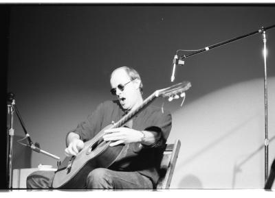 A medium-long shot of a seated guitar player, as he strums his guitar with a small metal tube. He is wearing a dark coloured shirt and sunglasses, and his jaw drops open. To the left side of the image is a microphone stand, and to right is a taller microphone stand.