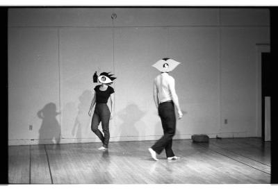 A black-and-white photo of Jane Ellison and Lola Ryan circling each other in the EDAM studio. They each wear a paper cutout on their heads evocative of an eye with large lashes and a pair of lips.