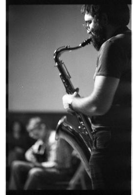 A man stands while playing saxophone. He is framed in the right half of the image. The background of the image is blurred, featuring an audience and guitar player. The image is in black and white.