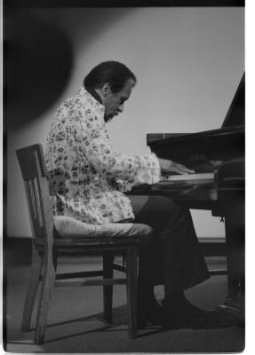 A long-shot of a man sitting in a wooden chair playing a grand piano. The image focuses on the man and the front of the grand piano. There is a spotlight framing him. His side-profile is featured and he is wearing a light-coloured patterned blazer, dark pants, and dark shoes.