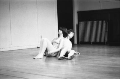 Two dancers are seated on the floor leans against each other. One of them has their knees propped up and reclines down onto the other dancer. The dance studio has wooden floors, walls with wainscoting, and a radiator in the back. 