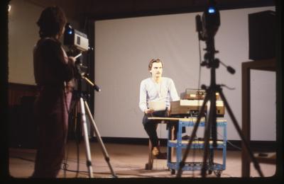 Dalibor Martinis sits on a stool in the Grand Luxe Hall in front of a projector screen, and next to recording equipment stacked on a wheeled cart. He wears a long sleeved shirt, jeans, brown shoes, and a microphone around his neck. Kate Craig is seen from behind operating a camera that’s pointed at him. 