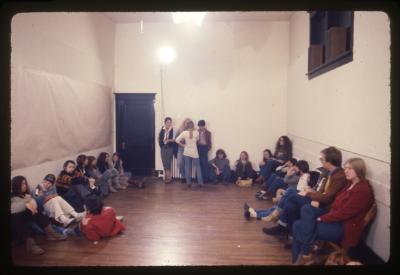 The audience during Sanja Iveković’s performance Meeting Points. Aside from a few folding chairs on the gallery’s periphery, the room is full of people sitting on the floor against the walls. The gallery door and window are painted black. 