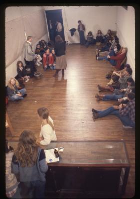 Seen from above, Sanja Iveković stands near a glass-top cabinet on the north end of the gallery. She wears a white long-sleeved shirt, and her hair is tied in a low ponytail. She looks out towards the audience, who is seated on the floor along the walls of the empty gallery. 