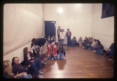 The audience during Sanja Iveković’s performance Meeting Points. Aside from a few folding chairs on the gallery’s periphery, the room is full of people sitting on the floor against the walls.

