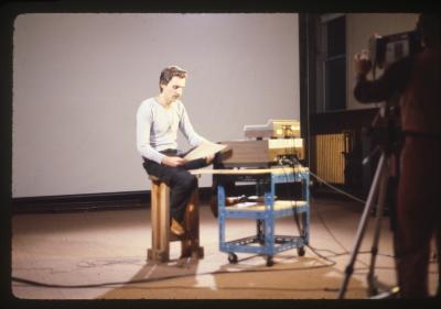 Dalibor Martinis sits on a wooden stool next to various recording equipment stacked on a wheeled cart. He reads from a piece of paper, and wears a white long sleeved shirt, jeans, and brown shoes. Kate Craig can be seen from behind operating a camera that is fixed on Dalibor. A projection screen is in the background.