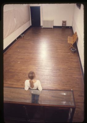 Seen from above, Sanja Iveković stands against a display case at the north end of the gallery at Western Front. She wears a white long sleeve shirt, jeans and her blonde hair is in a low ponytail. The room is empty aside from a set of three upholstered yellow folding chairs. 