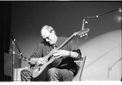 A medium-long shot of a seated guitar player, as he strums his guitar with a small metal tube. He is wearing a dark coloured shirt and sunglasses, and he hunches over his guitar smiling. To the left side of the image is a microphone stand, and to right is a taller microphone stand.