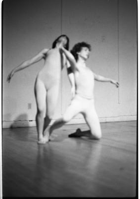 A dancer stretches their arms to either side of themselves with their knees bent and torso shifted to the right side of the image. The dancer next to them, kneels on the floor with one leg stretched back and their right arm extending to the other dancer's neck. Their other arm extends out to the right side of the image. 