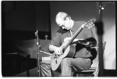 A medium-long shot of a seated guitar player, as he strums his guitar with a small metal tube. He is wearing a dark coloured shirt and sunglasses, and next to him is a microphone stand. The foreground has a blurred depiction of a piece of stage equipment.