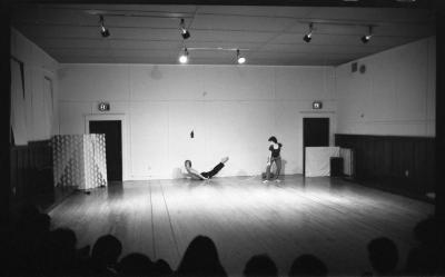 Lola Ryan lies on her stomach with her chest and legs lifted (as if in Locust pose). Jane Ellison stands behind her with a string tied around her waist that is connected to Lola’s wrist. The first row of the audience can be seen from behind.