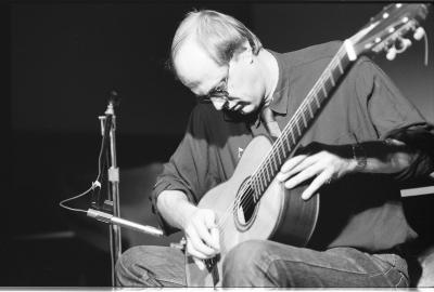 A medium-long shot of a seated guitar player, as he strums his guitar with a small metal tube. He is wearing a dark coloured shirt and sunglasses, and next to him is a microphone stand.