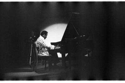 A man is performing as he plays the grand piano. There is a spotlight featured on him, and partially on the grand piano. There is a camera propped on a tripod behind the man. He is wearing a light-coloured patterned blazer and dark pants.