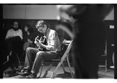 A man is sitting while playing guitar as an audience attends. The foreground is blurred, but features a man holding a saxophone.