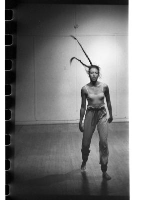 A woman steps forward in a dance studio. She is wearing a tank-top, long pants, and her hair is in two long braids. Her hair whips up into the air. The image is black and white.