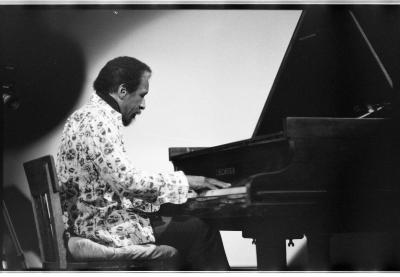 A medium-long shot of a man playing the grand piano. There is a spotlight on him. His side-profile is featured and he is wearing a light-coloured patterned blazer and dark pants.
