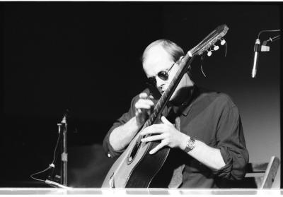 A medium-long shot of a seated guitar player, as he strums his guitar with a small metal tube. He is wearing a dark coloured shirt and sunglasses. Behind him are several pieces of stage equipment.
