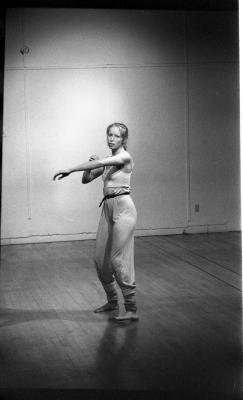 A woman stands in a dance studio with her arms extended out. Her body faces away from the camera. Her left arm is stretching forward and her right arm is bent and her hand touches her left elbow. Her head turns over her left shoulder. She is wearing a tank-top, long pants, and her hair is braided. The image is black and white.