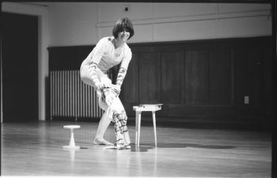 A dancer bends down and stretches their leg to put tights on, on their left side there is a small stool and on the right is a small table.