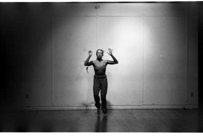 A woman stands in a dance studio with her right foot pointed down towards the floor. Her arms are bent and raised in the air. She is wearing a tank-top, long pants, and her hair is in two long braids. The image is black and white.