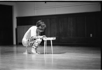 A dancer sits on a small stool, as they perch over a small table. The dance studio around them has wooden floors, walls with wainscoting, and a radiator in the back. 