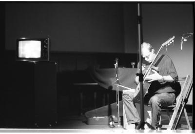 A long-shot of a seated guitar player, as he strums his guitar with a small metal tube. He is wearing a dark coloured shirt and sunglasses. Behind him are several pieces of stage equipment, and to the left of the image is a small CRT television on top of a cabinet. The foreground has blurred depictions of stage equipment. 