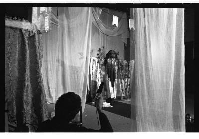 Curtains and a banner hang from the ceiling. There is a tarp runway that rests on the floor. Centred between some of these curtains is a sculpture. In the foreground, a man, seen from behind, holds a microphone with a raised arm. 
