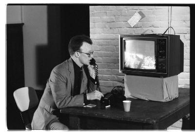 Mike Haslam sits at a wooden desk and speaks into a touch-tone phone. Next to him, a television monitor shows two men. Haslam wears a suit and glasses. 