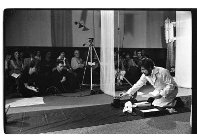 A man crouches on a tarp runway and in front of him are various objects. Two curtains drape down from the ceiling. The background features an audience and in front of them is a camera hoisted on a tripod. The image is in black and white.
