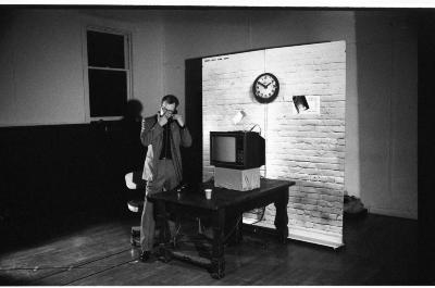 Mike Haslam stands at a wood table stacked with a television set. He is dressed in a suit and holds a telephone receiver up to his ear while he pushes his glasses up at the bridge of his nose. There is a free-standing backdrop behind him printed with a clock hanging on a brick wall. 