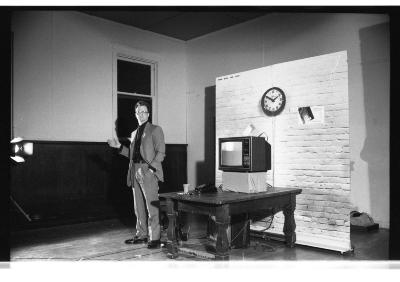 Mike Haslam stands in the Grand Luxe Hall with one hand in the pocket of his suit pants. Next to him, a television monitor is stacked on a wooden table that is positioned in front of a backdrop of a brick wall.