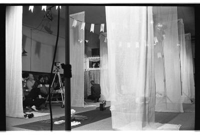 A tarp runway is layed on the ground with various objects on top of it. Six curtains flow down from the ceiling, three on either side of the tarp. The background features an audience with a camera hoisted on a tripod. Two people crouch down between the curtains. There are four banners that loom over this set. This image is in black and white.
