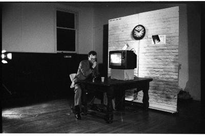 Mike Haslam sits at a wooden table and speaks into a touch-tone phone next to a television monitor. A backdrop of a brick wall stands freely behind the TV.